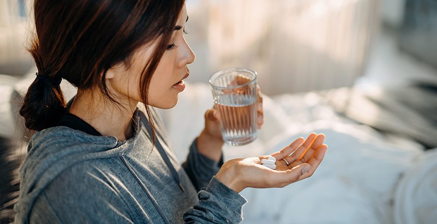 Jeune femme asiatique assise sur un lit tenant des médicaments dans la main et un verre d'eau