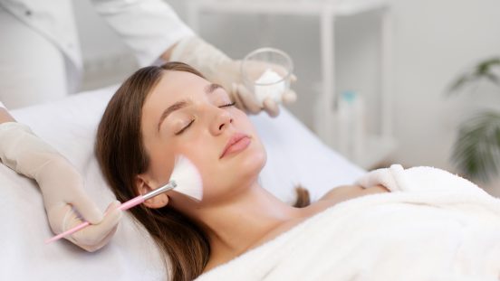 Une femme bénéficie d'un soin du visage professionnel allongée sur une table; le chirurgien esthétique lui applique un peeling sur le visage